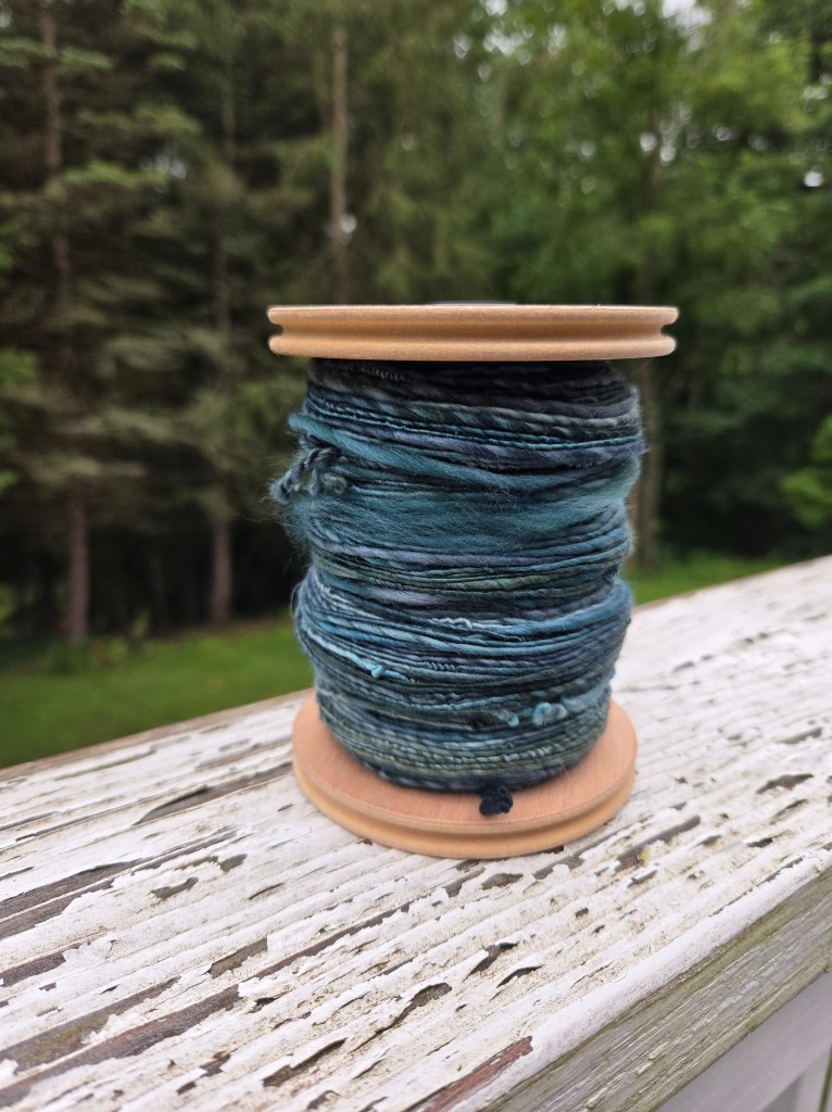 a bobbin of green-blue yarn, lumpily spun, against a background of green trees