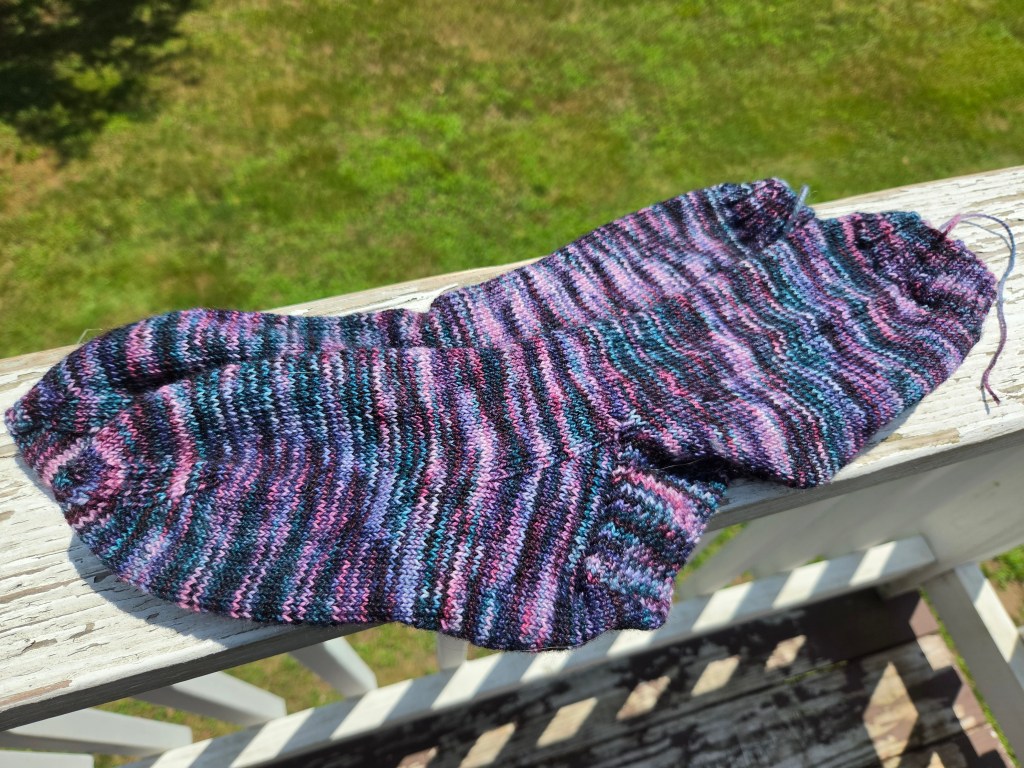 a pair of handknit socks, on a white porch railing; they are very colorful, in shades of pink, teal, dark purple, and black